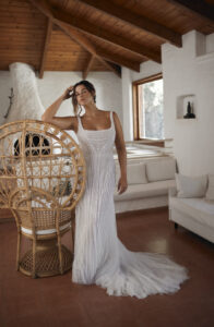 Dana Harel Jolene Wedding Dress from a bridal shop in Chicago, IL. The model has her arm on a chair and is posing in a room with a wood-burning fireplace.