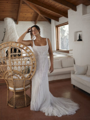 Dana Harel Jolene Wedding Dress from a bridal shop in Chicago, IL. The model has her arm on a chair and is posing in a room with a wood-burning fireplace.