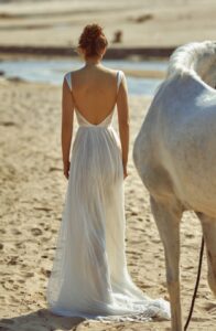 A model is looking backward next to a horse in the sand wearing a Nara by Dana Harel wedding dress from a bridal shop in Chicago, IL.