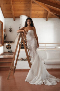 Dana Harel Venice Wedding Dress from a Bridal Shop in Chicago, IL. The model is posing with her arm on a ladder and three steps in the background.