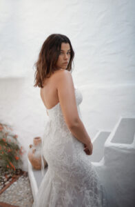 Dana Harel Venice Wedding Dress from a Bridal Shop in Chicago, IL. The model is posing in front of a white stucco stairwell showing the side and back of the dress.