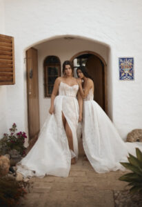 Dana Harel Kim wedding dress from a bridal shop in Chicago, IL. Two models are sitting standing in front of an arched exterior doorway.