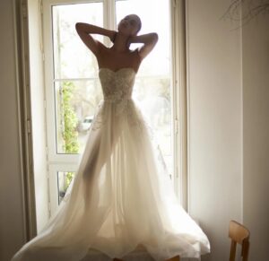 A model wearing a Dana Harel Peony wedding dress backlit by a window. The dress is a whimsical A-line gown with a tulle skirt and a delicate floral applique and can be found at a bridal shop in Chicago, IL