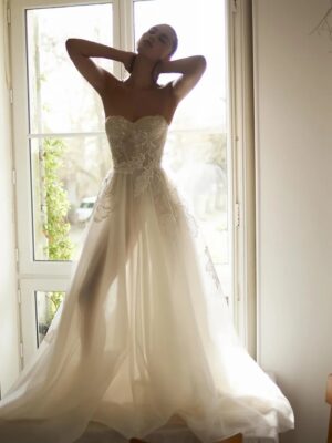 A model wearing a Dana Harel Peony wedding dress backlit by a window. The dress is a whimsical A-line gown with a tulle skirt and a delicate floral applique and can be found at a bridal shop in Chicago, IL