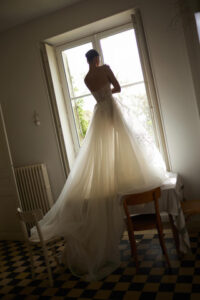 A model wearing a Dana Harel Peony wedding dress backlit by a window. The dress is a whimsical A-line gown with a tulle skirt and a delicate floral applique and can be found at a bridal shop in Chicago, IL