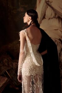 A model is showing the back of her delicate mini wedding dress "Bonvolio" by designer Kim Kassas. This dress is has sheer embroidered fabric draped across one shoulder. It's available through Mira Couture in Chicago, IL