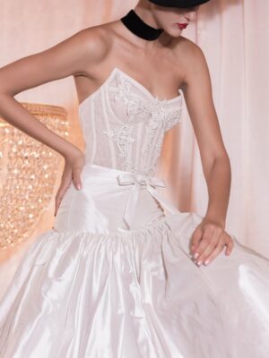 A model wearing the tea length Ella by Kim Kassas wedding dress, featuring a corset and bow in the front. Available at a bridal shop in Chicago, IL