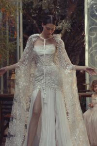 A model is wearing a strapless lace corset wedding dress "Giovanna" by designer Kim Kassas and a dramatic cape. This dress has a fit and flare style and is available through Mira Couture in Chicago, IL