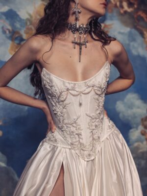 A model is wearing Kim Kassas's Isabelle strapless wedding dress with a high slit and detailed bodice with large religious jewelry