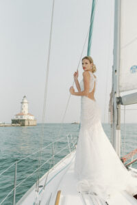 A model alone stands with her back to the camera on a sail boat. The eye-catching fit and flare wedding dress features a plunge neckline and beautifully draped pearl beading. The dress is available at bridal shop Mira Couture in Chicago, IL.