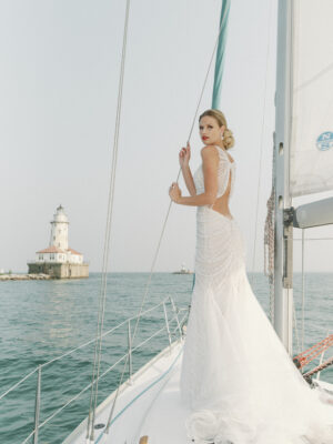 A model alone stands with her back to the camera on a sail boat. The eye-catching fit and flare wedding dress features a plunge neckline and beautifully draped pearl beading. The dress is available at bridal shop Mira Couture in Chicago, IL.