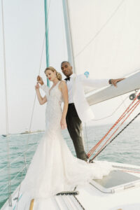 A model and her groom stand on a sail boat in Lake Michigan in Chicago, IL. The eye-catching fit and flare wedding dress features a plunge neckline and beautifully draped pearl beading. The dress is available at bridal shop Mira Couture in Chicago, IL.