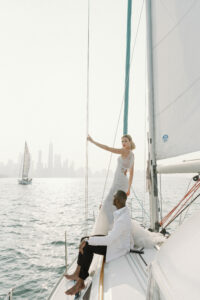 A model and her groom stand on a sail boat in Lake Michigan with the Chicago skyline in the background. The eye-catching fit and flare wedding dress features a plunge neckline and beautifully draped pearl beading. The dress is available at bridal shop Mira Couture in Chicago, IL.