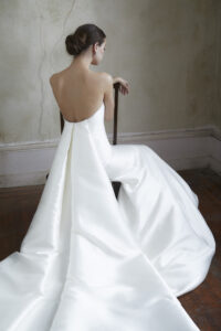 Back view of a model sitting and wearing a clean fit and flare gown featuring a classy crescent neckline and a detachable train from a bridal shop in Chicago, IL.