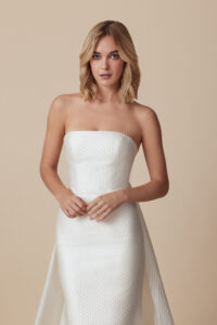 A mid-ground frontal view of a model wearing Bobbi by Ti Adora Allison Webb, a fit and flare gown that features unique geometric embossed sateen fabric, crescent neckline, and a detachable train from a bridal shop in Chicago, IL.