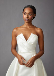 Mid-ground front view of a model wearing a trumpet style gown that features a pleated bodice, and detachable open overskirt from a bridal shop in Chicago, IL.