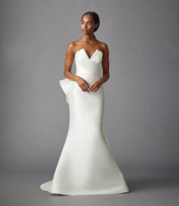 Full front view of a model wearing a trumpet style gown that features a pleated bodice, detachable open overskirt, and a beautiful asymmetrical bow from a bridal shop in Chicago, IL.