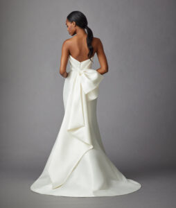 Back view of a model wearing a trumpet style gown that features a pleated bodice, detachable open overskirt, and a beautiful asymmetrical bow from a bridal shop in Chicago, IL.