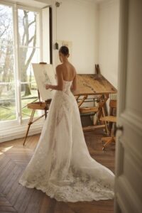 A bride stands in front of a drawing board in her Moe by Dana Harel wedding gown. It is an elegant fitted lace wedding dress made of sautage lace. Featuring a high collar neck and a classy open back.