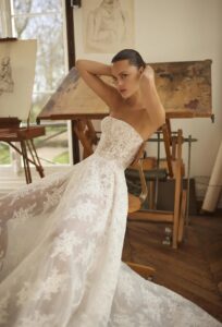 A leans back in a chair in her Moe by Dana Harel wedding gown. It is an elegant fitted lace wedding dress made of sautage lace. Featuring a high collar neck and a classy open back.