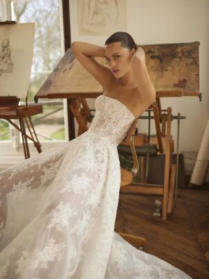 A leans back in a chair in her Moe by Dana Harel wedding gown. It is an elegant fitted lace wedding dress made of sautage lace. Featuring a high collar neck and a classy open back.