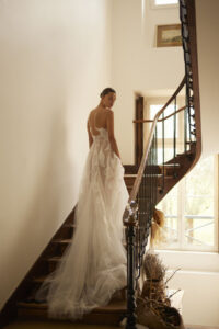 A bride shows the back of her Lily by Dana Harel bridal gown. It is a feminine soft A-line wedding dress made out of a beautiful tulle skirt. It features delicate Calla Lily flowers cascading from the bodice down the train, creating an illusion-like painting on the body.