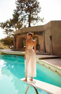 A bride shows the back of her stunning Tulip by Dana Harel wedding dress. It is a floral strapless corset gown that features 3D motifs, intricate lace detailing, and a gathered tea-length skirt.