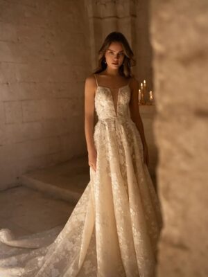 A model in a chapel faces the camera as she shows off Helga, by Eva Lendel, an elegant and timeless A-line gown. Featuring delicate lace applique, a deep v-neckline, and a stunning skirt that cascades into a beautiful long train. Adorned with a dainty bow tied at the waist.