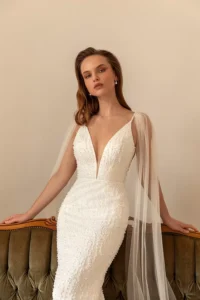 A closeup of a model leaning on the top of a sofa while wearing the bold and glamorous beaded fitted gown Perla by Eva Lendel that features a plunge neckline and an elegant open back.