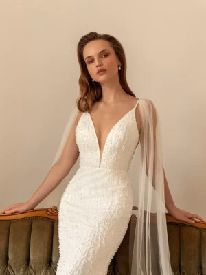 A closeup of a model leaning on the top of a sofa while wearing the bold and glamorous beaded fitted gown Perla by Eva Lendel that features a plunge neckline and an elegant open back.