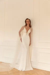 A model leans against a wall while wearing the bold and glamorous beaded fitted gown Perla by Eva Lendel that features a plunge neckline and an elegant open back.