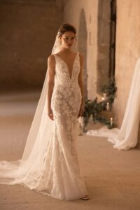 A model waits at the end of an aisle in a church wearing a veil and the Santiago by Eva Lendel wedding dress from Mira Couture. It is an elegant and whimsical fitted gown that features a deep v-neckline and is adorned with delicate 3D floral applique.