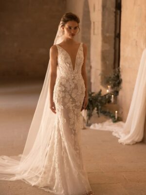 A model waits at the end of an aisle in a church wearing a veil and the Santiago by Eva Lendel wedding dress from Mira Couture. It is an elegant and whimsical fitted gown that features a deep v-neckline and is adorned with delicate 3D floral applique.