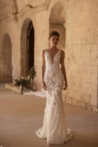 A model looking at the camera in a church wearing the Santiago by Eva Lendel wedding dress from Mira Couture. It is an elegant and whimsical fitted gown that features a deep v-neckline and is adorned with delicate 3D floral applique.