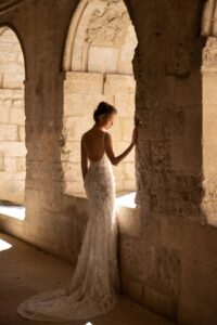 A model looking outside of a church window wearing the Santiago by Eva Lendel wedding dress from Mira Couture. It is an elegant and whimsical fitted gown that features a deep v-neckline and adorned with delicate 3D floral applique.