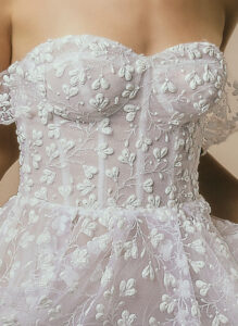 A closeup of the bust of the Denise by Neta Dover wedding dress, an alluring sweetheart corset gown that features dainty 3D floral applique. It's completed with romantic off-shoulder draped sleeves and is available at a bridal shop in Chicago, IL.