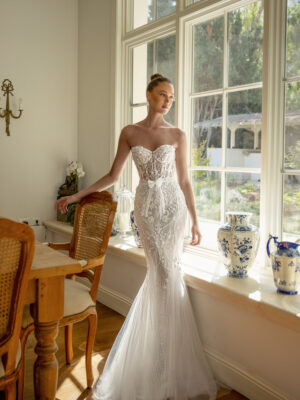 A bride looks out of a window while wearing the Patreece by Neta Dover wedding dress. It is a strapless fitted bridal gown featuring a corset top, intricate beaded lace, and a sweetheart neckline. It's available at a bridal shop in Chicago, IL.