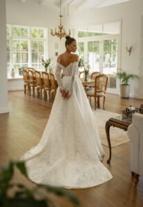 A bride shows the back of the Pier by Neta Dover wedding dress. It's a romantic Chantilly lace ballgown featuring a sweetheart neckline and charming lace long sleeves. It's available at a bridal shop in Chicago, IL.