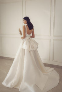 A bride stands and shows the back of the Renee by Neta Dover wedding dress. It is a classy A-line bridal gown adorned with an oversized bow. It's available at a bridal shop in Chicago, IL.