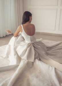 A bride sits while wearing the Renee by Neta Dover wedding dress. It is a classy A-line bridal gown adorned in 3D florals and an elegant square neckline with an oversized bow. It's available at a bridal shop in Chicago, IL.