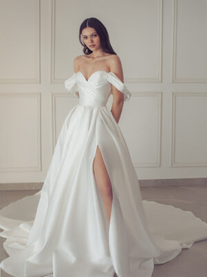 A bride wears the Rona by Neta Dover wedding dress. It is an A-line silk Mikado bridal gown that features a pleated sweetheart neckline, leg slit, and an elegant long train. It is available at a bridal shop in Chicago, IL.