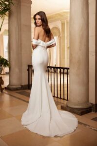 A model shows the back of the 5061 by Paloma Blanca wedding dress, an elegant and timeless crêpe fitted bridal gown with a removable pleated off-shoulder portrait collar and a sleek long train. It is available at a bridal shop in Chicago, IL.