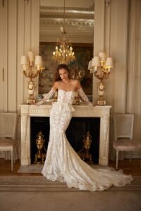 A model wears a floral lace, fitted wedding dress with long sleeves, a sweetheart neckline, and a ruffle peplum belt available at a bridal shop in Chicago, IL.