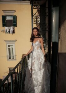 A bride shows the front of her wedding dress, Chiara by Eden Aharon. This dreamy A-line bridal gown features dainty straps that cascade into a sheer corset bodice. It has large floral embellishments across the bodice and full skirt. See it in Chicago, IL.