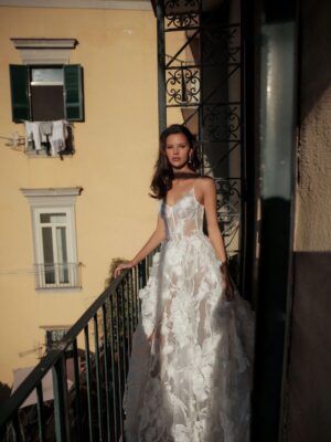 A bride shows the front of her wedding dress, Chiara by Eden Aharon. This dreamy A-line bridal gown features dainty straps that cascade into a sheer corset bodice. It has large floral embellishments across the bodice and full skirt. See it in Chicago, IL.