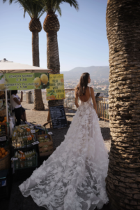 A bride shows the back of her wedding dress, Chiara by Eden Aharon. This dreamy A-line bridal gown features dainty straps that cascade into a sheer corset bodice. It has large floral embellishments across the bodice and full skirt. See it in Chicago, IL.