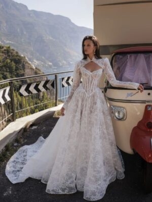 A bride shows the front of her wedding dress, Denia by Eden Aharon. This bridal gown is embellished with delicate lace and a dropped Basque waistline. It's paired with a sheer corset bodice and a detachable high-neck bolero. See it in Chicago, IL.