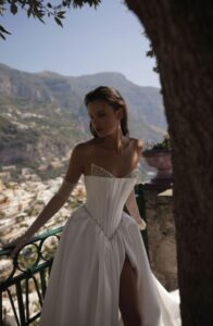 A bride shows the bodice of her Micol by Eden Aharon wedding dress. This corset bodice is adorned with a dropped Basque waistline. It’s made of silk Mikado fabric and features a slit. See it in Chicago, IL.