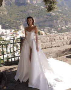 A bride shows a full view of the front of her Micol by Eden Aharon wedding dress. This strapless dress features a corset bodice that is adorned with a dropped Basque waistline. It’s made of silk Mikado fabric and features a slit. See it in Chicago, IL.