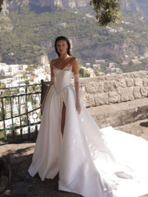 A bride shows a full view of the front of her Micol by Eden Aharon wedding dress. This strapless dress features a corset bodice that is adorned with a dropped Basque waistline. It’s made of silk Mikado fabric and features a slit. See it in Chicago, IL.